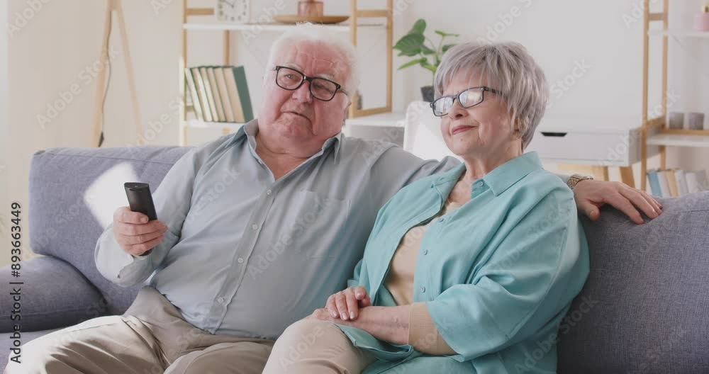 Elderly senior couple using remote control to change channel watching TV sitting on sofa at home. Wife and husband enjoying television show resting on couch. Leisure in retirement concept. 4k video. Elderly senior couple using remote control to change channel watching TV sitting on sofa at home. Wife and husband enjoying television show resting on couch. Leisure in retirement concept. 4k video.