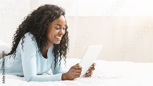 Interesting content. Millennial african girl reading blog on digital tablet lying in bed, free space