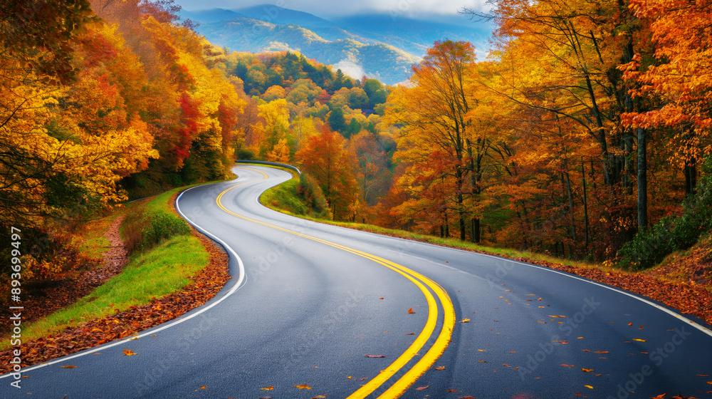 Fototapeta premium Scenic curved road through a forest with vibrant autumn foliage in yellow, orange, and red hues. Wet road and misty mountains in the background.