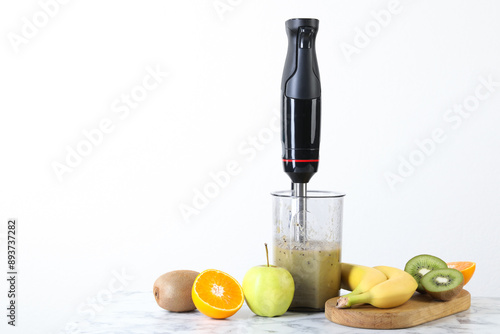 Fototapeta Naklejka Na Ścianę i Meble -  Hand blender with mixture of ingredients and fresh fruits on white marble table, space for text
