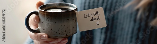 A close-up of a hand holding a rustic mug with a message tag that reads 'Let's talk about it,' inviting a conversation over coffee.