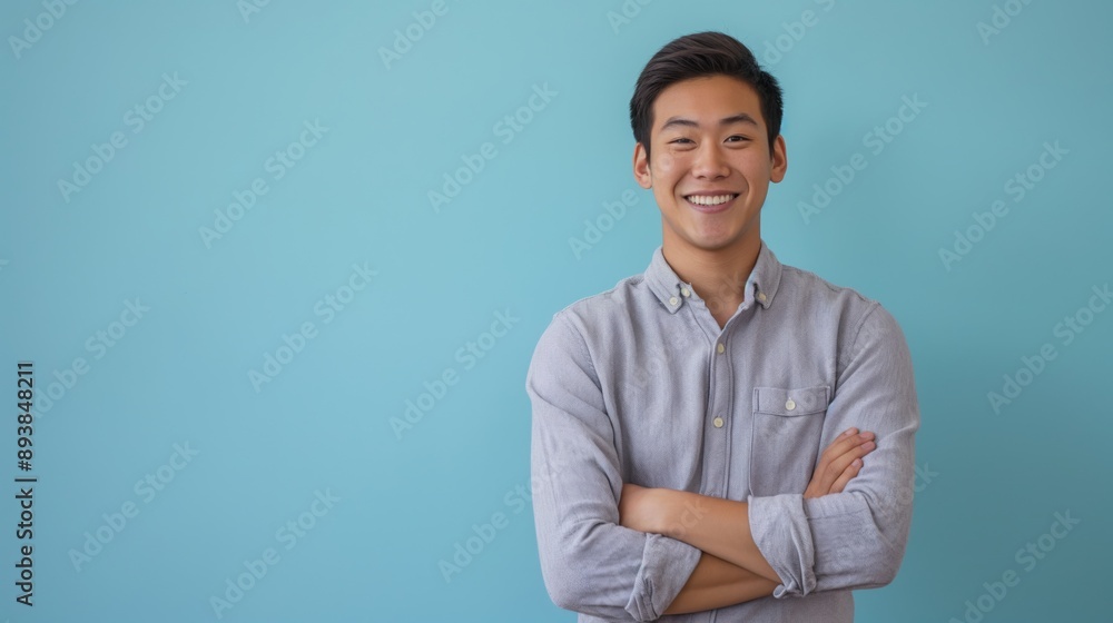Confident Young East Asian Man Smiling with Arms Crossed Against Blue Background for Professional Use Confident Young East Asian Man Smiling with Arms Crossed Against Blue Background for Professional Use