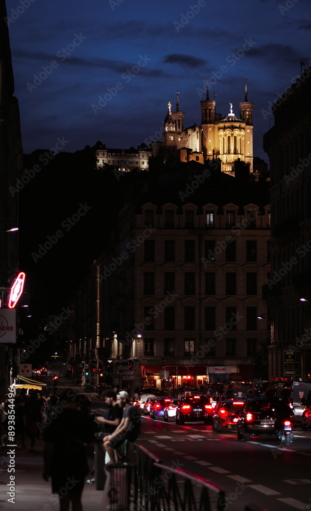 Obraz premium Lyon, France - August 12 2023. People stroll along the lively streets at night, illuminated by lanterns and shop signs, with the Basilica Notre Dame de Fourvière standing majestically in the backgroun