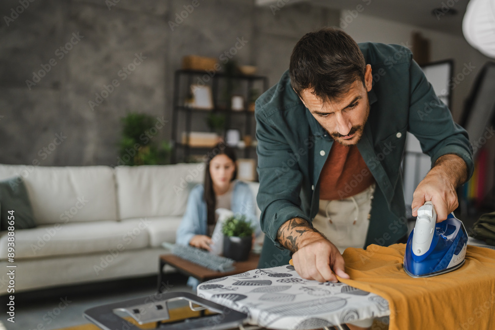 Adult man iron the wardrobe on the iron board while woman clean dust Adult man iron the wardrobe on the iron board while woman clean dust