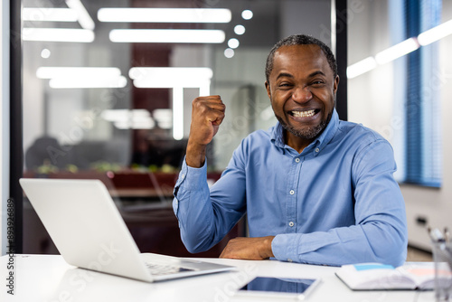 Excited businessman celebrating success at office desk with laptop, raised fist and big smile. Professional environment, natural light, and happy emotions indicating achievement and joy.