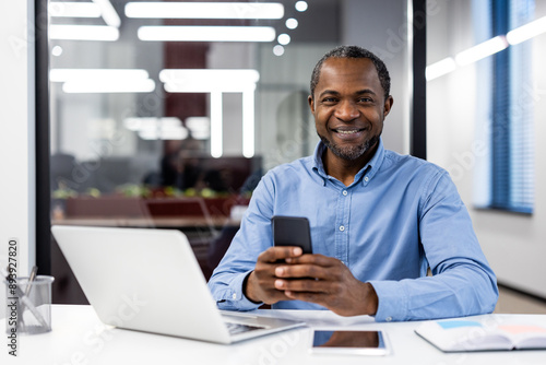 Confident businessman smiling while using smartphone at modern office desk. Professional workspace with laptop, tablet, and office supplies. Concept of business, technology, and corporate lifestyle.