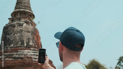 Man tourist taking picture of ancient pagoda with smartphone in Ayutthaya historical park, Thailand. Male traveler photographing istorical Buddhist stupa with mobile phone on archaelogical site photo