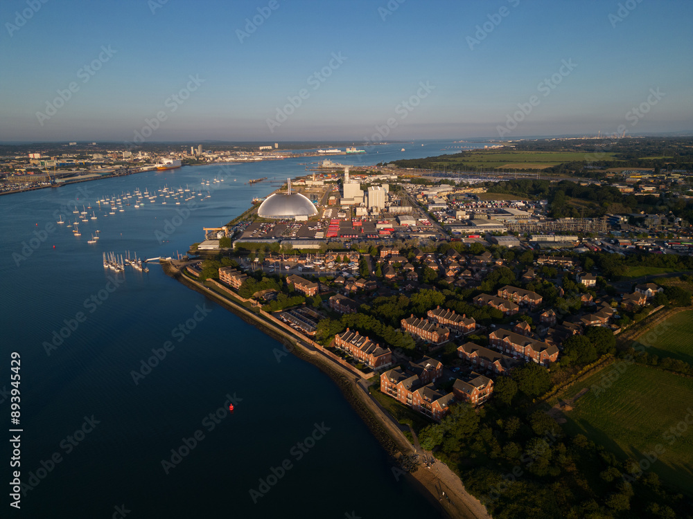 Fototapeta premium Marchwood waterfront residential area and Power Station at industrial estate. Aerial view of Southampton harbor and the city at distance. Sailing boats mooring. Calm summer day with beautiful sunset.