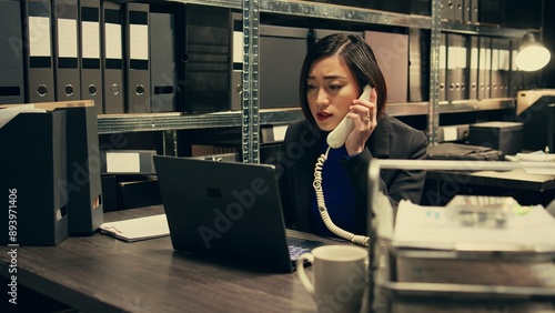 Law officer verifying details for the private investigation, working in archive room. Detective cross checks information on landline phone call, contacting people from records. Camera B. photo