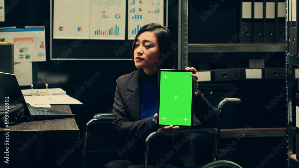 Law agent in wheelchair shows greenscreen display on tablet, using isolated mockup layout on device in disability friendly archive room. Investigator holding gadget with chromakey screen. Camera A. Law agent in wheelchair shows greenscreen display on tablet, using isolated mockup layout on device in disability friendly archive room. Investigator holding gadget with chromakey screen. Camera A.