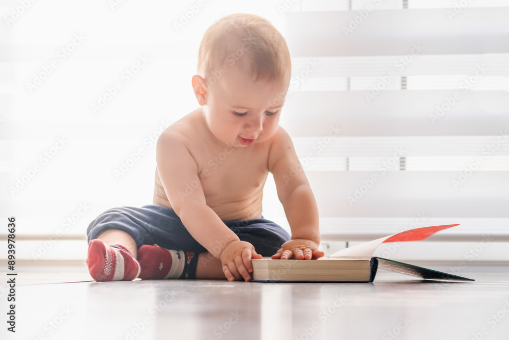 Child development concept. Funny portrait of sweet baby boy sitting on the floor at home and reading book. Horizontal image. Child development concept. Funny portrait of sweet baby boy sitting on the floor at home and reading book. Horizontal image.