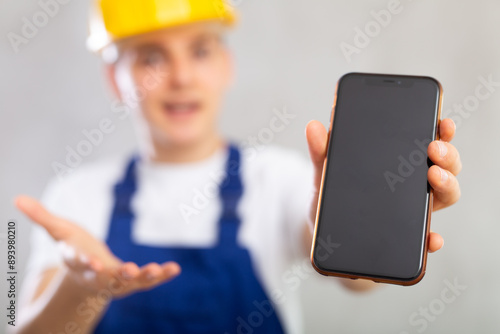 Surprised young man worker demonstrating screen of mobile phone with astonishment standing against background of light unicolored wall photo