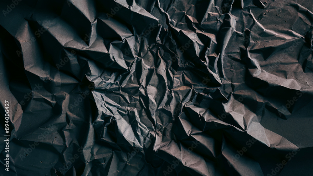 A close-up view of crumpled, wrinkled fabric. The fabric appears dark, possibly black or deep gray, and is densely packed with numerous folds and creases. A close-up view of crumpled, wrinkled fabric. The fabric appears dark, possibly black or deep gray, and is densely packed with numerous folds and creases.