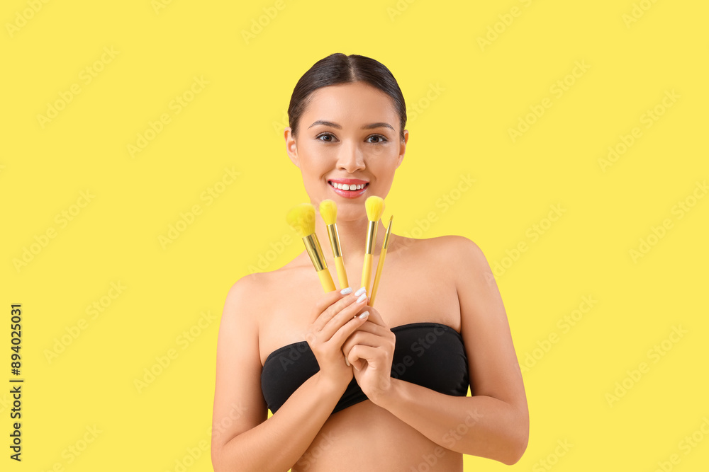 Young Asian woman with makeup brushes on yellow background Young Asian woman with makeup brushes on yellow background