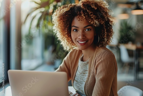 A cheerful woman with curly hair smiles while working on her laptop in a modern office space, enjoying a productive moment.