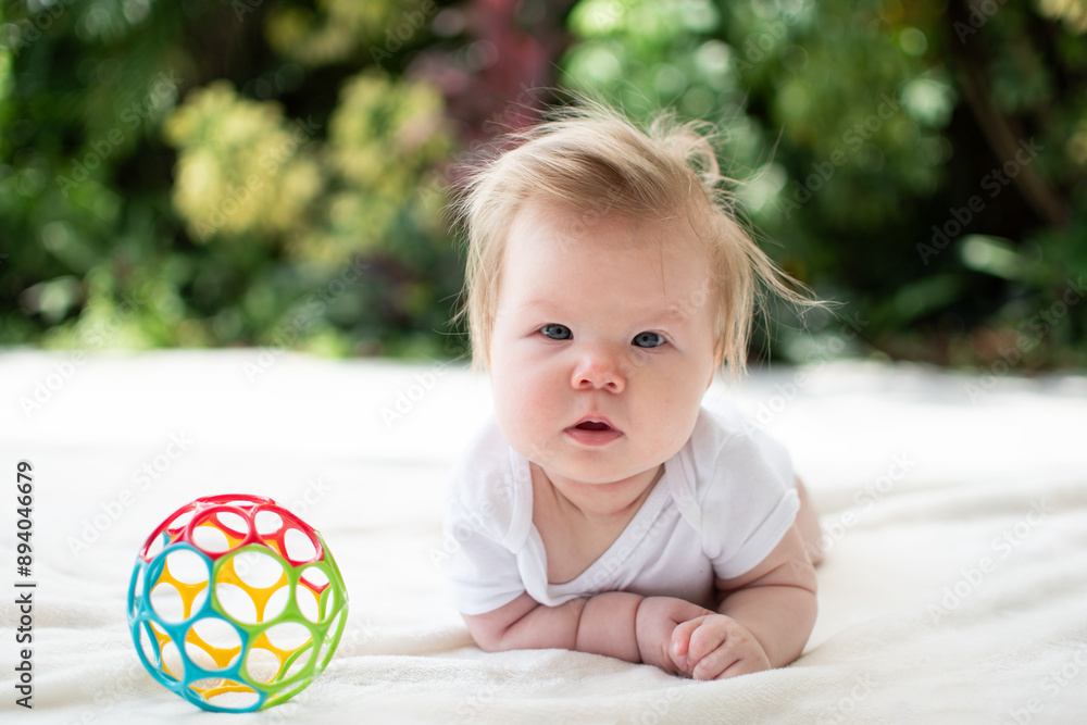 Cute Caucasian baby girl during tummy time looking at the camera. 3-month-old baby playing on the floor with a colorful ball