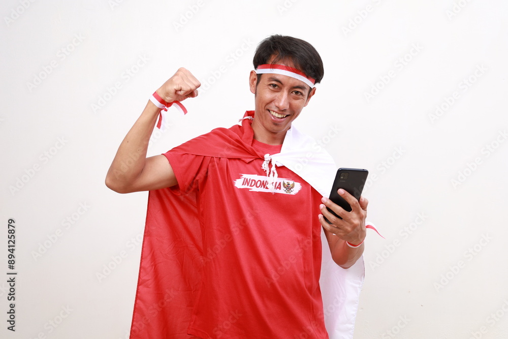 Excited Asian young boy with red white ribbon for celebrating Indonesia independence day standing holding cell phone while clenching hand