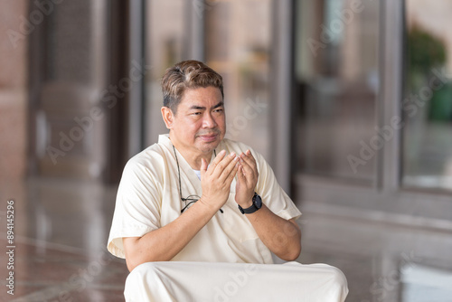 Fifty-something Muslim Malay person in religious attire, wearing a white outfit and black hat, praying devoutly in a wide glass-enclosed marble area outside very large mosque in Putrajaya, Malaysia. photo