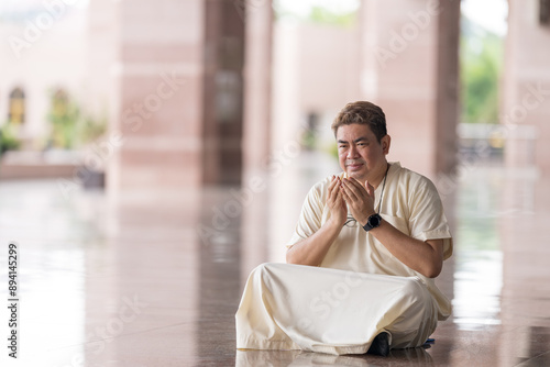 Fifty-something Muslim Malay person in religious attire, wearing a white outfit and black hat, praying devoutly in a wide glass-enclosed marble area outside very large mosque in Putrajaya, Malaysia. photo