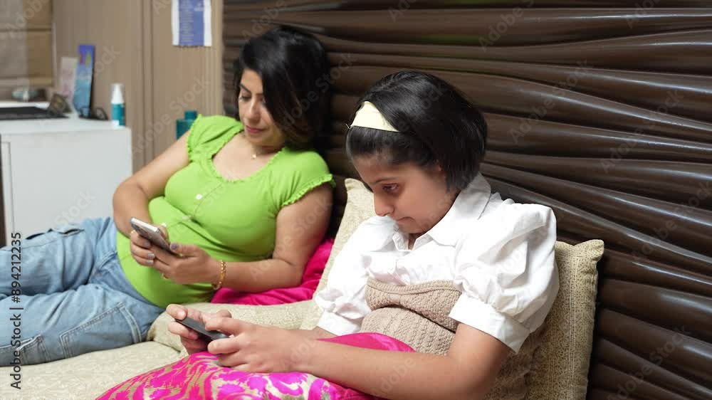 Young indian mother and little daughter using smart phone while lying on bed at home. People addicted to mobile smart phone, playing games, watch videos online, Children's screen addiction,Technology Young indian mother and little daughter using smart phone while lying on bed at home. People addicted to mobile smart phone, playing games, watch videos online, Children's screen addiction,Technology