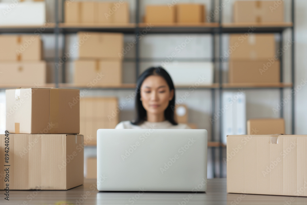 A woman is working on a laptop surrounded by boxes, likely managing inventory in a warehouse or preparing orders for an online store. A woman is working on a laptop surrounded by boxes, likely managing inventory in a warehouse or preparing orders for an online store.