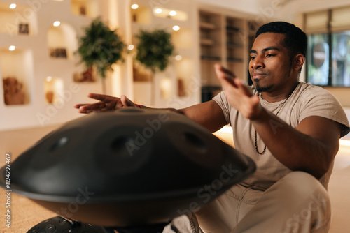 Portrait of serene black male in trance practicing native music traditional drumming instrument playing with hands sitting on floor in lotus pose. Relaxed African man drumming rhythm on tank drum photo