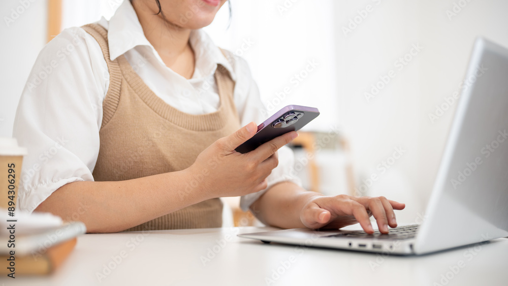 Fototapeta premium An Asian woman working remotely from a cafe, multitasking, using her smartphone and her laptop.