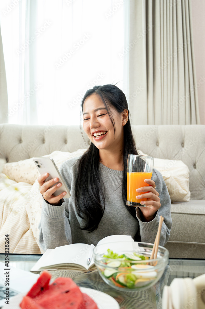 An Asian woman is laughing while reading chat on her phone and having breakfast at a coffee table. An Asian woman is laughing while reading chat on her phone and having breakfast at a coffee table.