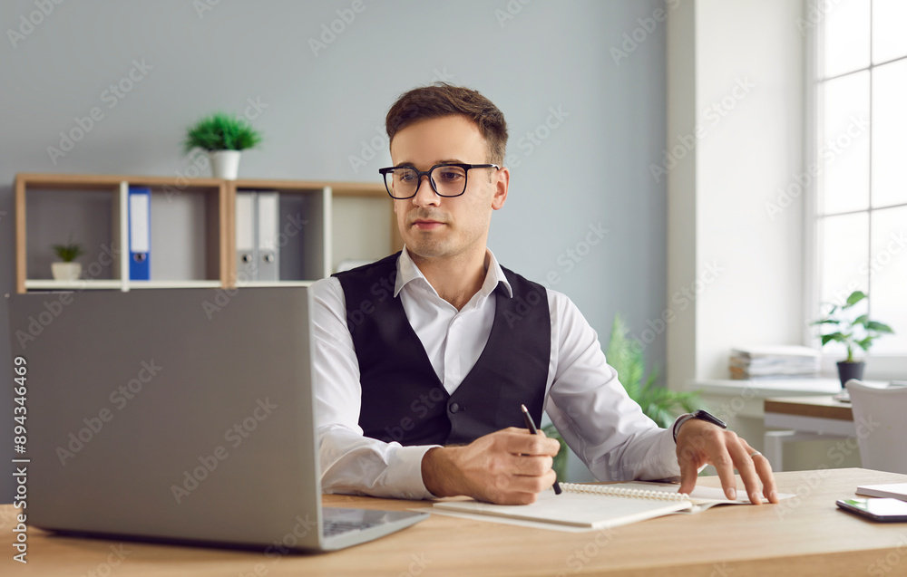 Portrait of young confident business man sitting on workplace at the desk working on a laptop at office making notes or planning daily meeting and appointment looking at computer monitor screen. Portrait of young confident business man sitting on workplace at the desk working on a laptop at office making notes or planning daily meeting and appointment looking at computer monitor screen.