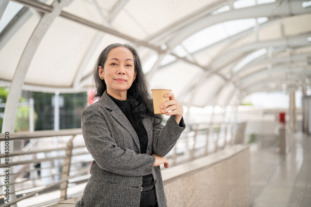 A confident mature Asian businesswoman is smiling at the camera while standing on a skywalk. A confident mature Asian businesswoman is smiling at the camera while standing on a skywalk.