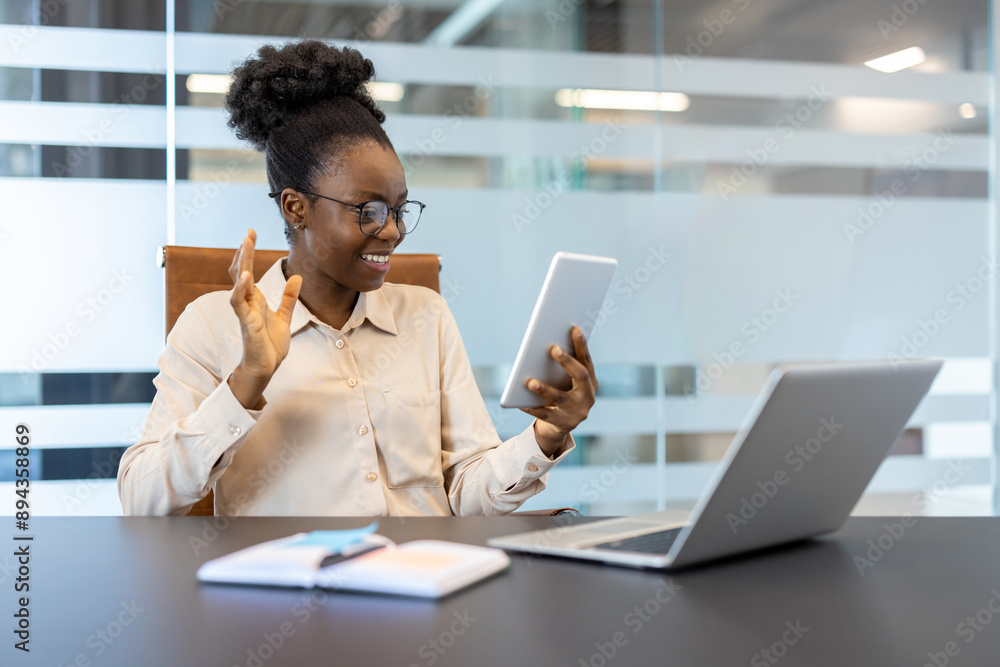 Smiling businesswoman waving during video call on tablet in modern office setting with laptop and notebook on desk. Engaged in online meeting conveying communication, connection, and technology. Smiling businesswoman waving during video call on tablet in modern office setting with laptop and notebook on desk. Engaged in online meeting conveying communication, connection, and technology.