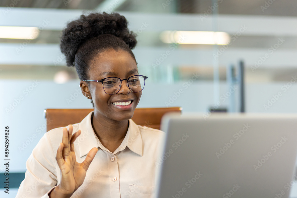 Smiling woman wearing glasses participating in video call, waving hand, sitting in modern office. Professional setting with soft focus background. Business communication, remote work concept. Smiling woman wearing glasses participating in video call, waving hand, sitting in modern office. Professional setting with soft focus background. Business communication, remote work concept.