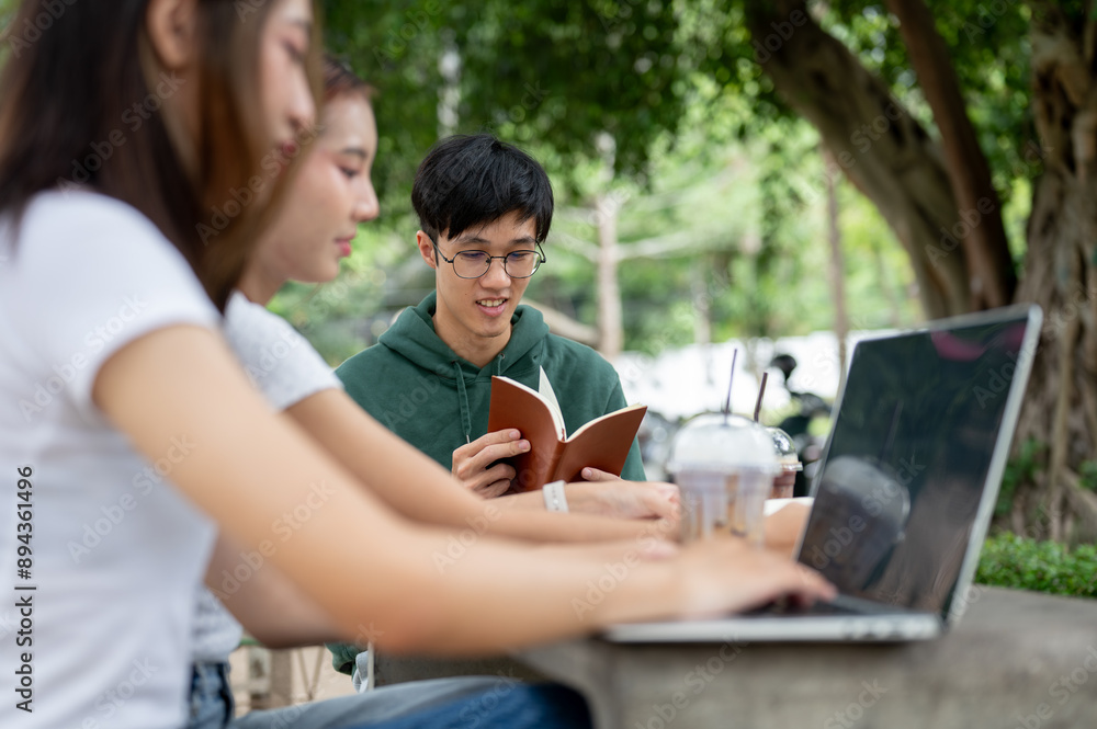 A young Asian male student is reading a book while working with his friends at an outdoor table. A young Asian male student is reading a book while working with his friends at an outdoor table.
