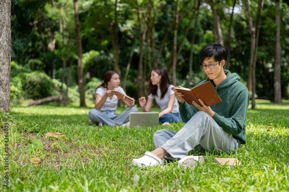 A smart, nerdy young Asian male college student is focused on reading a book in the campus park. A smart, nerdy young Asian male college student is focused on reading a book in the campus park.