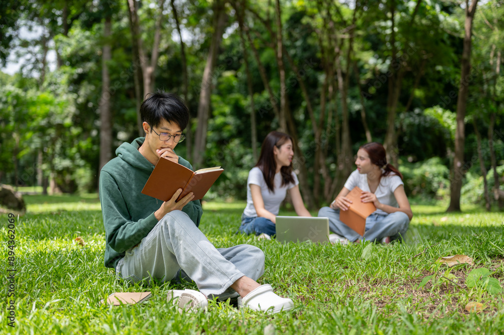 A smart, nerdy young Asian male college student is focused on reading a book in the campus park. A smart, nerdy young Asian male college student is focused on reading a book in the campus park.
