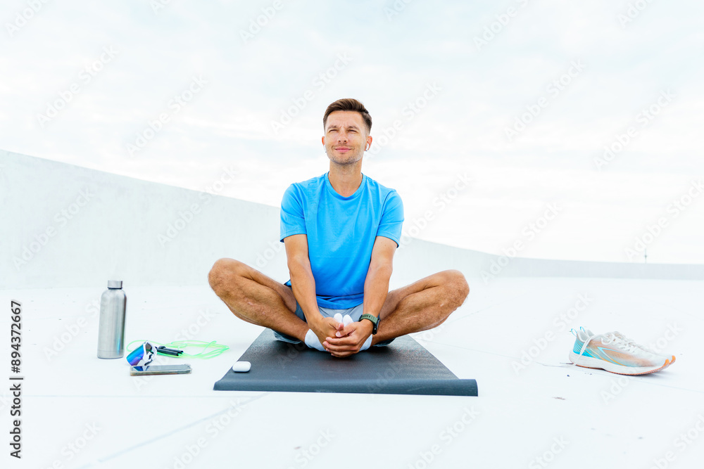 Fototapeta premium A man sits cross-legged on a mat, embracing a tranquil moment after exercise, surrounded by fitness gear.