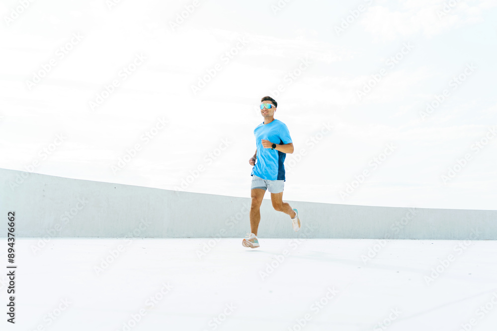 Man in a blue outfit runs outdoors, wearing sunglasses and a smartwatch, with a bright sky above. Man in a blue outfit runs outdoors, wearing sunglasses and a smartwatch, with a bright sky above.