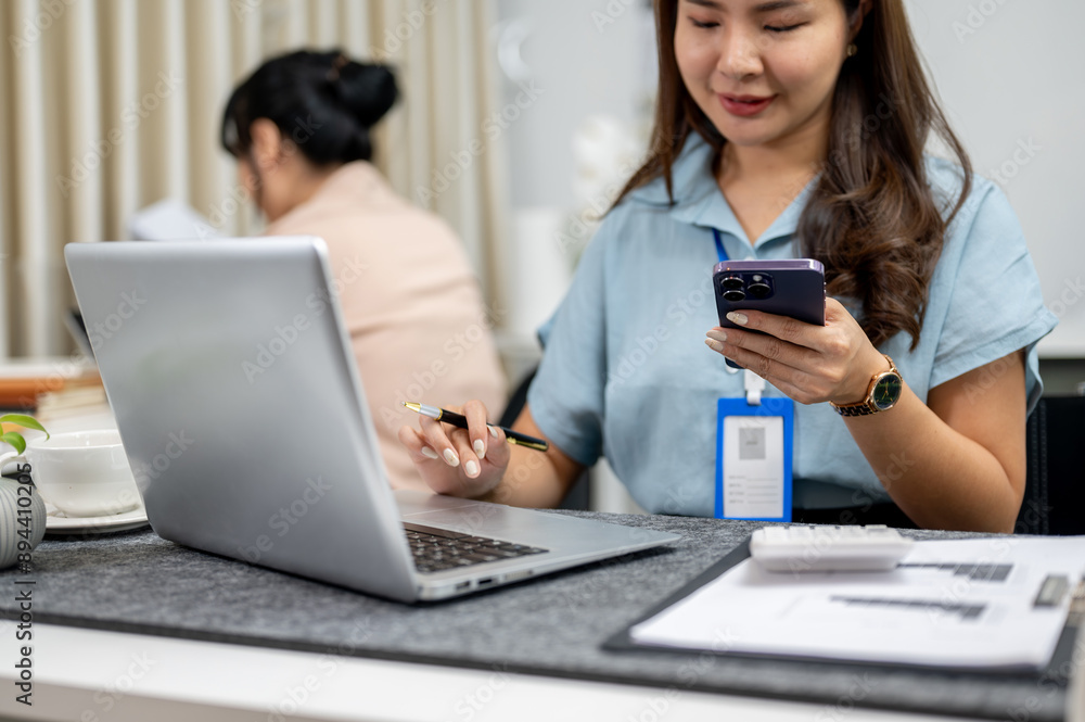 A positive Asian female office employee is using her smartphone while working on her laptop. A positive Asian female office employee is using her smartphone while working on her laptop.
