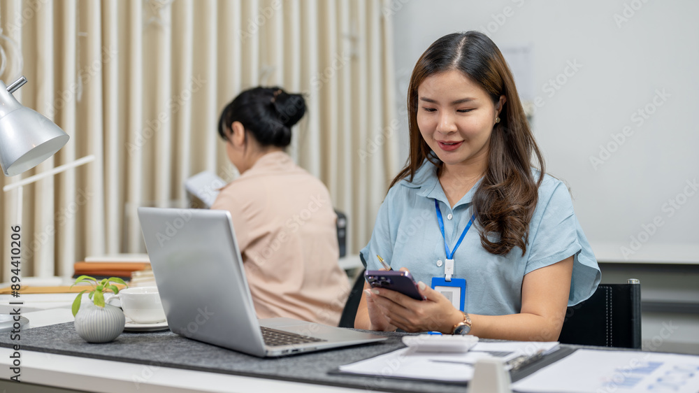 A positive Asian female employee using her smartphone at her desk in the office, checking messages. A positive Asian female employee using her smartphone at her desk in the office, checking messages.