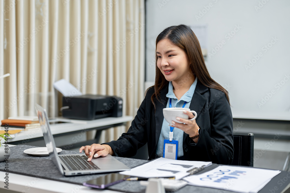 A happy Asian businesswoman is having coffee while working on her laptop computer at her desk. A happy Asian businesswoman is having coffee while working on her laptop computer at her desk.
