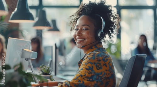 Young, smiling mixed-race girl contact center agent answering calls with a headset. An office desk-bound Hispanic businesswoman with a curly afro on a desktop computer taking a call. photo