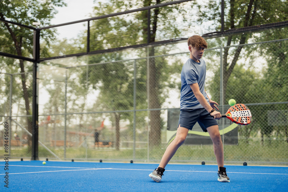 Obraz premium Focused young male player hitting the ball during a game of padel tennis on an outdoor blue court.