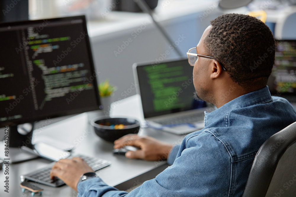 Back view of male African American IT engineer focused on writing code looking at computer screen on blurred background, copy space Back view of male African American IT engineer focused on writing code looking at computer screen on blurred background, copy space