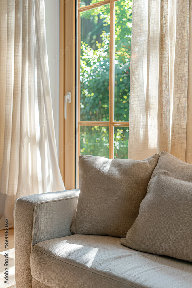 Fototapeta premium Close-up of a sofa in a living room with a window and curtains. The couch has a beige linen fabric. Light colors are used. Sunlight is coming through the trees on this sunny day.