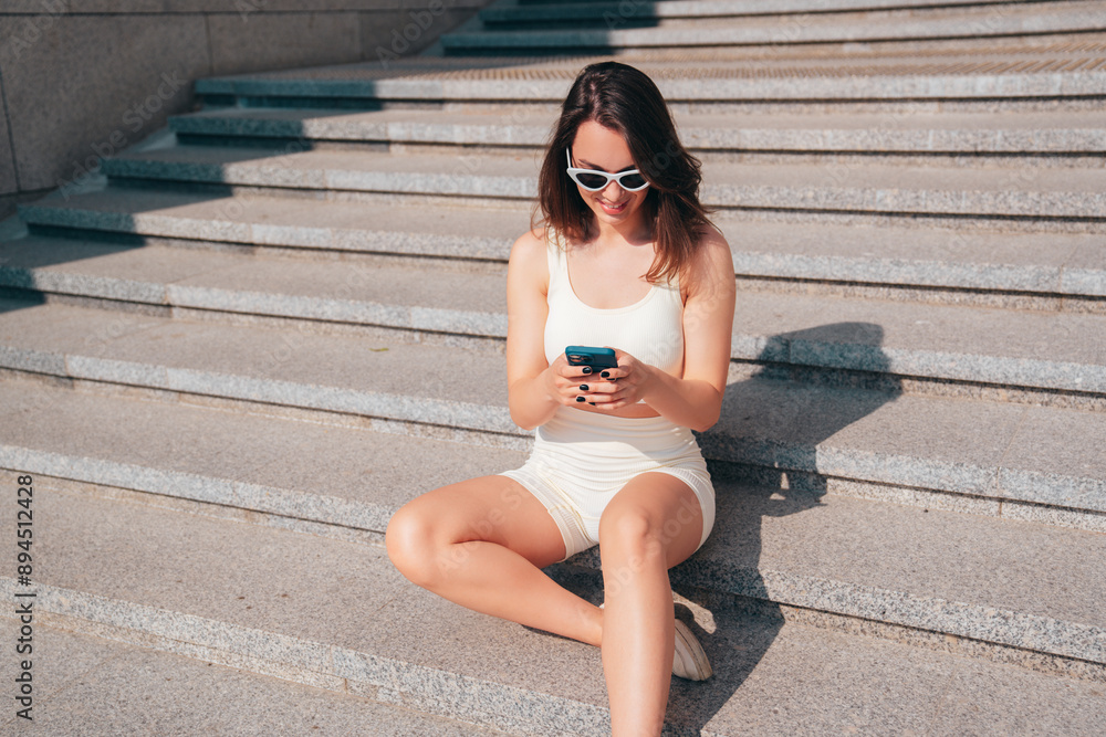 Obraz premium Young beautiful smiling female in trendy summer white cycling shorts and tank top clothes. Sexy carefree woman posing in street. holds smartphone, uses mobile apps, looks at phone screen. Texting sms