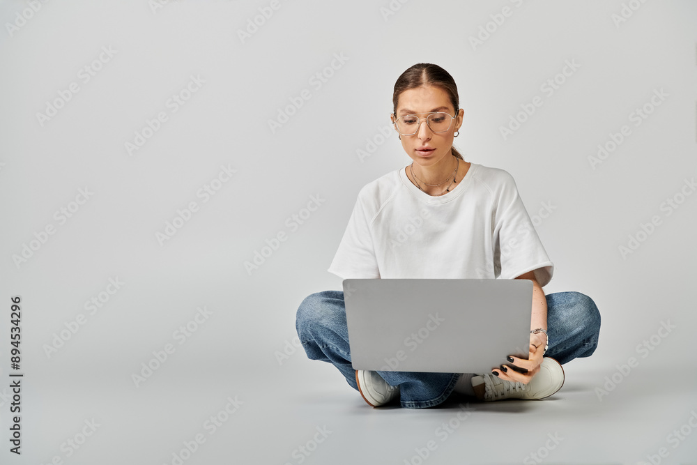 A young woman in a white t-shirt and glasses sitting on the floor, engrossed in her laptop on a grey background. A young woman in a white t-shirt and glasses sitting on the floor, engrossed in her laptop on a grey background.