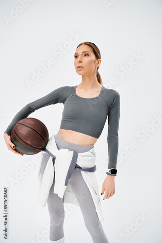 A sporty young woman in active wear confidently holds a basketball in her right hand against a grey background. photo