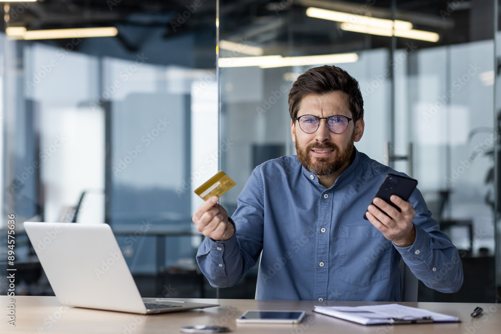 Frustrated man holding credit card and smartphone in office Frustrated man holding credit card and smartphone in office
