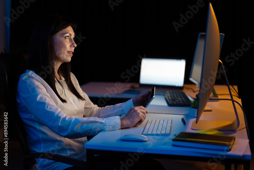 Woman Working Late At Office, Using Computer And Smartphone, Focused And Engaged In Her Tasks photo