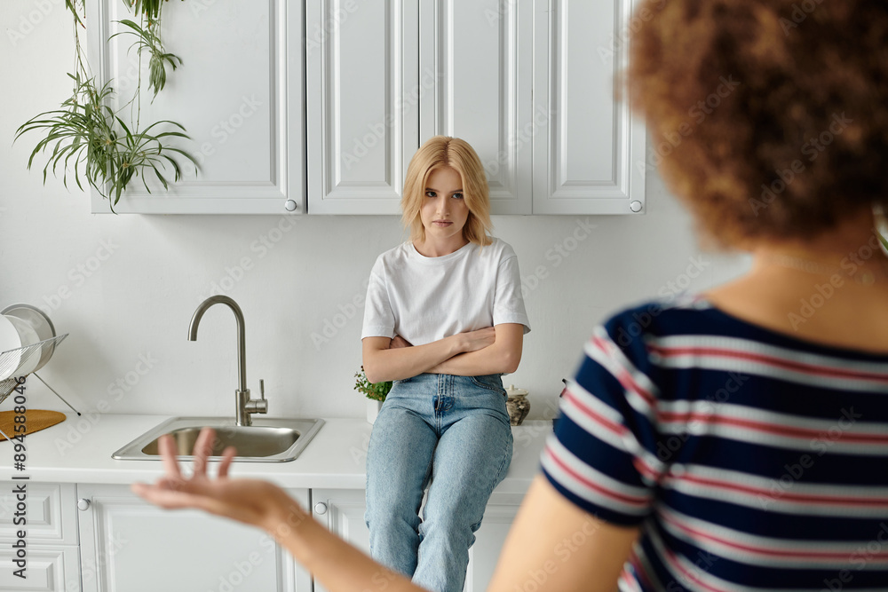 © Bliss - Two women stand in a kitchen, one with her arms crossed, the other gesturing with a displeased expression. © Bliss - Two women stand in a kitchen, one with her arms crossed, the other gesturing with a displeased expression.